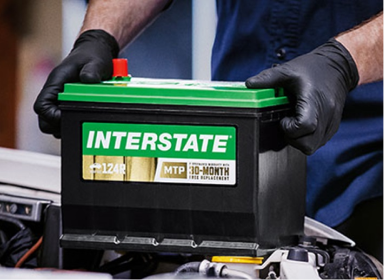 Technician wearing blue button-up and black rubber gloves grabbing onto a green and black Interstate car battery product by the sides.