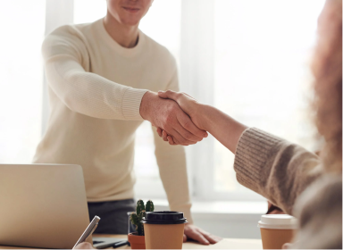 Person in a white sweater shaking hands with a person in a grey sweater. The room is located in an office space with white walls and tall windows.