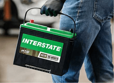 Technician wearing blue denim jeans and black rubber gloves holding a green and black Interstate car battery by the handle inside an auto shop.