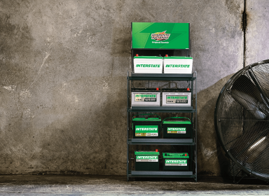 Store display rack of Interstate Battery products standing in front of a cement wall with a giant fan to its right.