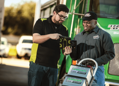 Interstate deliveryman with a dolly full of batteries speaking with a store manager while both are looking at a tablet. Both are standing outdoors and outside of the Interstate delivery truck.