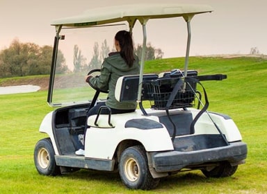 Person with a ponytail, wearing a green jacket, black pants, and white shoes is driving a golf cart over a golf course.