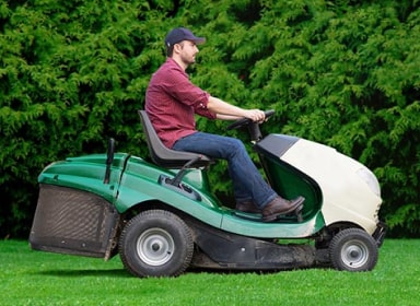 Man with facial hair, a red button-up, blue jeans and brown shoes riding a green and white seated lawn mower in fornt of green trees.