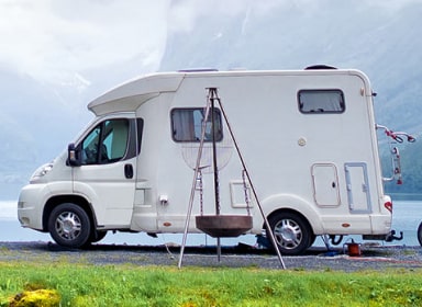 White RV parked in front of a lake and mountains. In front of the RV is a hanging fire pit.