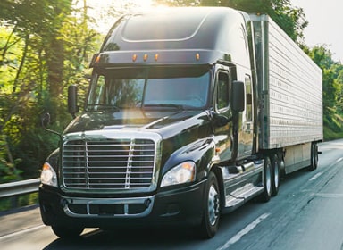 Black Semi-Truck driving on right side of the road next to green trees with sunlight shining down.